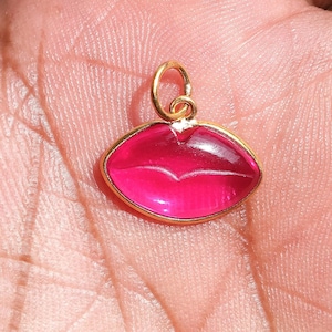 May include: A gold-colored pendant in the shape of a pair of lips, filled with a vibrant pink material. The pendant has a small loop at the top for attaching to a chain or bracelet. The lips are outlined in gold.