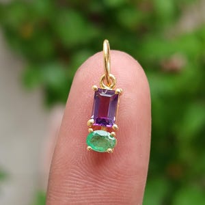May include: A gold pendant featuring a rectangular purple amethyst and an oval green emerald. The gemstones are set in a gold frame with a small loop at the top for a chain. The pendant is held up by a finger.