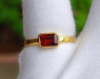 May include: A gold ring with a rectangular, deep red gemstone. The gemstone is set in a gold bezel, and the band is polished gold. The ring is shown against a white background.