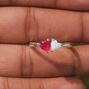 May include: A silver ring featuring two heart-shaped gemstones. One gemstone is a vibrant red, and the other is a shimmering white. The ring is set against a blurred background, highlighting the jewelry's design.