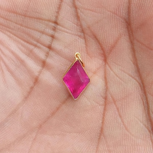 Puede incluir: Un pequeño colgante dorado con una gema rosa vibrante en forma de diamante. El colgante está suspendido de un pequeño lazo en la parte superior. La gema tiene múltiples facetas, que reflejan la luz.