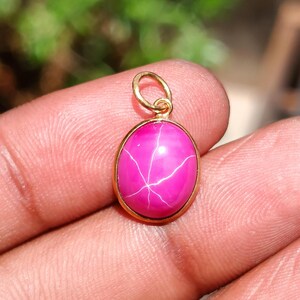 Könnte beinhalten: Ein kleiner ovaler Anhänger mit einem leuchtend pinkfarbenen Cabochon-Stein, auf dessen Oberfläche ein deutliches sechsstrahliges Sternmuster zu sehen ist. Der Stein ist in eine polierte goldfarbene Metallfassung eingefasst und hat einen kleinen goldenen Ring.