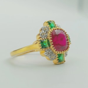 May include: A gold ring featuring a large, oval-cut ruby center stone, surrounded by smaller green and white accent stones. The ring has a detailed, ornate design with a polished finish, showcasing a classic and elegant style.