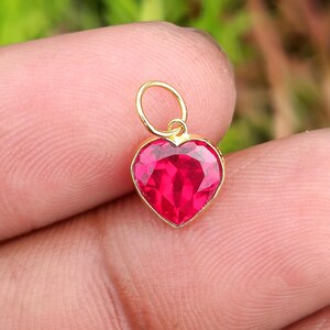May include: A small heart-shaped pendant featuring a vibrant, faceted red gemstone set in a polished gold-toned metal frame. The charm includes a circular gold jump ring at the top, designed for attachment to a necklace chain or bracelet link.