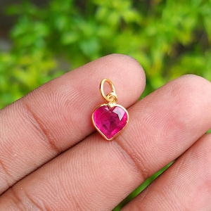 May include: A gold heart pendant with a bright pink gemstone. The pendant has a small loop at the top for a chain. The gemstone is faceted, catching the light. The background is a blurred green.