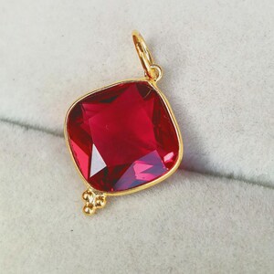 May include: A gold-colored pendant with a large, square-cut, deep red gemstone. The gemstone is faceted, catching the light. The pendant has a small loop at the top for a chain and a cluster of small gold beads at the bottom.