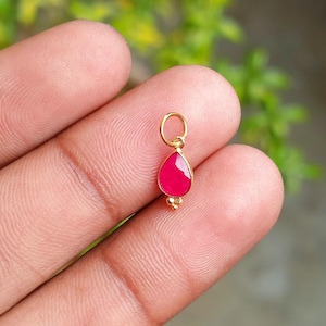 May include: A small, gold pendant featuring a teardrop-shaped, bright pink gemstone. The pendant has a loop for a chain and two small gold beads at the base. The background is a blurred green.