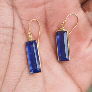 May include: A pair of gold-colored earrings with rectangular, deep blue gemstones. The gemstones are set in a gold frame and have a polished, reflective surface. The earrings have a simple, elegant design with a hook closure.