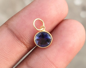 Puede incluir: Un colgante dorado con una piedra preciosa redonda de color azul oscuro. La piedra preciosa está engastada en un bisel dorado y tiene una superficie facetada. El colgante tiene un pequeño lazo en la parte superior para sujetarlo a una cadena. El colgante está en una mano.