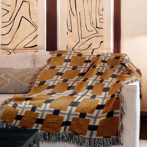 May include: A woven throw blanket featuring a repeating grid pattern in shades of mustard yellow, charcoal gray, and light gray, with fringed edges draped over a light-colored sofa next to a metallic lamp on a black square side table.