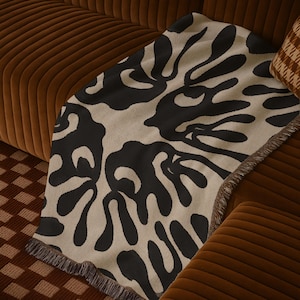 May include: A rectangular woven throw blanket featuring a high-contrast pattern of large, organic black shapes against a cream-colored background. The blanket has a textured knit appearance and is finished with short, dark fringe along the edges.