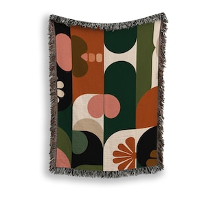 May include: A woven throw blanket with a geometric pattern in shades of rust, green, black, and cream. The design features abstract shapes, including circles and arches. The blanket has a fringed edge.