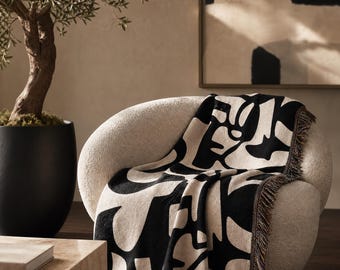 May include: A woven throw blanket featuring a high-contrast black and cream abstract organic pattern. The rectangular textile has a soft texture and is finished with short, dark brown fringe along the edges, draped over a rounded cream chair.