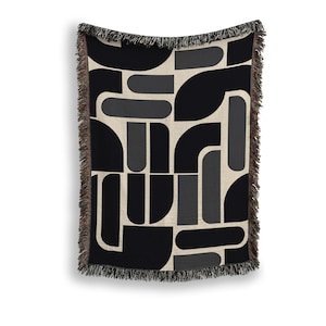 May include: A woven throw blanket with a geometric pattern in black, gray, and beige. The design features abstract shapes and lines. The blanket has a fringed edge.