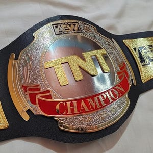 Puede incluir: Un cinturón de campeonato de lucha libre con una correa de cuero negro. La placa central es plateada con detalles dorados y las letras "TNT" en oro. Una pancarta roja dice "CHAMPION". El logo de AEW es visible.
