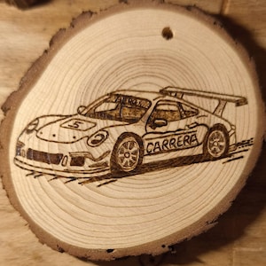 Puede incluir: Un adorno de rodaja de madera circular con un diseño de pirograbado de un coche de carreras. El coche se muestra de perfil con el número 5 en el capó y la palabra CARRERA impresa en el lateral. La veta natural de la madera es visible y tiene un pequeño agujero perforado cerca del borde superior.