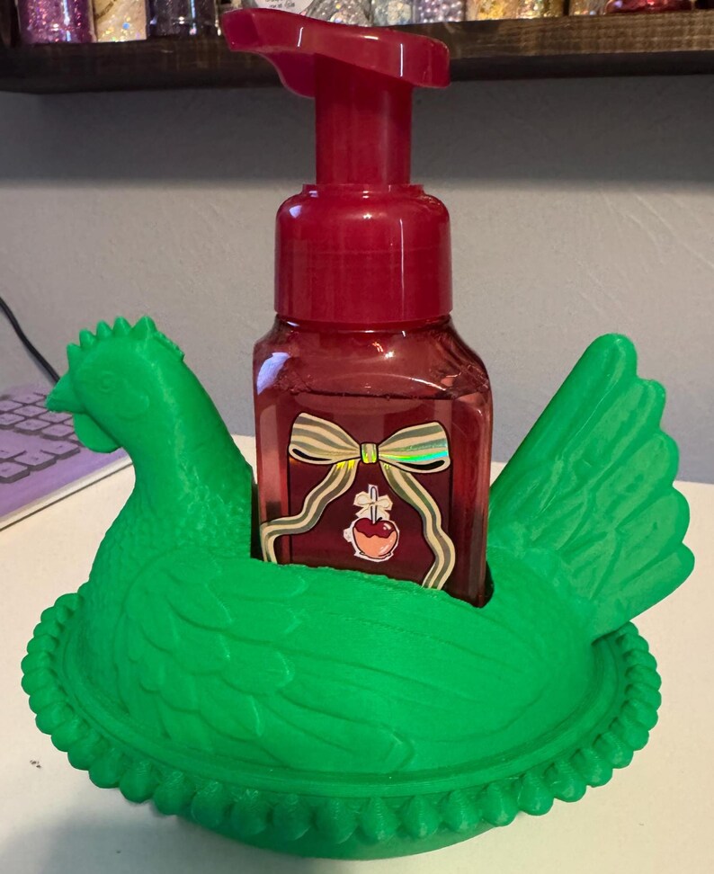 Puede incluir: Un dispensador de jab&oacute;n rojo con un dise&ntilde;o de lazo se encuentra dentro de un soporte con forma de pollo verde brillante. El soporte tiene un dise&ntilde;o detallado de un pollo con una base en forma de nido. El dispensador tiene una bomba roja y una botella transparente.