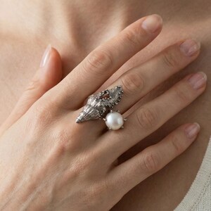 Puede incluir: Anillo plateado con diseño de concha y una perla grande y redonda. El anillo se lleva en un dedo, destacando sus intrincados detalles y su estética inspirada en la naturaleza. El diseño del anillo incluye pequeños acentos oscuros.