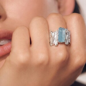 Könnte beinhalten: Ein silberner Ring mit einem rechteckigen, hellblauen Edelstein. Der Ring hat ein strukturiertes, rindenartiges Design um den Edelstein. Der Ring befindet sich an einem Finger.