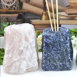 Può includere: Due porta diffusori a bastoncino in pietra naturale. Uno è di colore rosa chiaro, l'altro blu scuro con un motivo maculato. Il supporto blu contiene diversi bastoncini di legno. Le pietre hanno una superficie ruvida e strutturata.