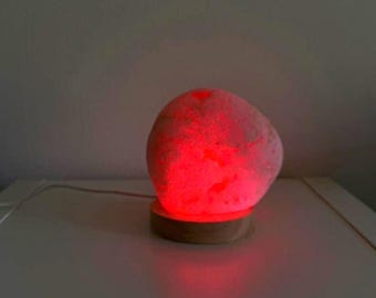 May include: A pink, illuminated rock lamp. The lamp has a smooth, rounded shape and sits on a small, round wooden base. The light emitted is a vibrant red, creating a warm glow. The lamp is plugged in with a white cord.