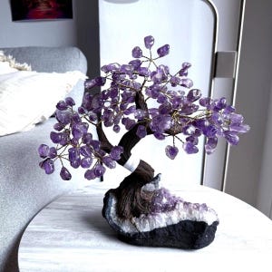 May include: A decorative amethyst tree sculpture. The tree features a dark brown trunk and branches adorned with purple amethyst stones. The base is a dark rock with a white and purple crystal formation.