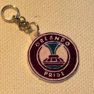 May include: A circular keychain displaying the Orlando Pride logo. The logo is a stylized design in white, teal, and purple, with the words "ORLANDO" and "PRIDE" in white lettering. The keychain has a silver-coloured metal ring and chain.