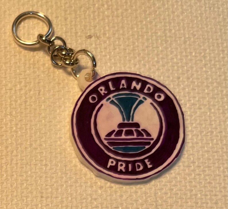 May include: A circular keychain displaying the Orlando Pride logo. The logo is a stylized design in white, teal, and purple, with the words "ORLANDO" and "PRIDE" in white lettering. The keychain has a silver-coloured metal ring and chain.