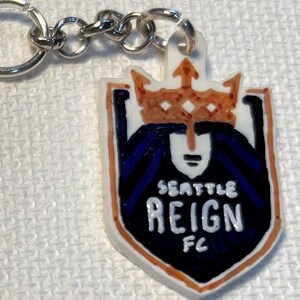May include: A keychain with the Seattle Reign FC logo. The logo is a shield shape with a white background, a crown, and the words "SEATTLE REIGN FC". The keychain has a silver chain.