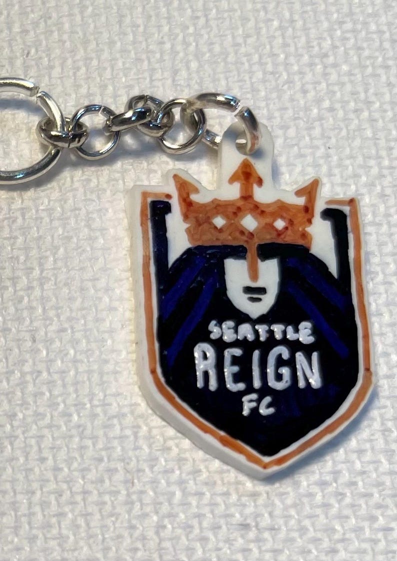 May include: A keychain with the Seattle Reign FC logo. The logo is a shield shape with a white background, a crown, and the words "SEATTLE REIGN FC". The keychain has a silver chain.