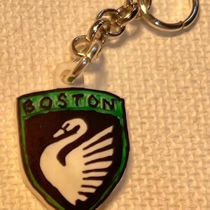 May include: A keychain with a shield-shaped emblem displaying the word "BOSTON" in green. The emblem features a black background and a white swan design. The keychain has a silver-coloured chain and ring.