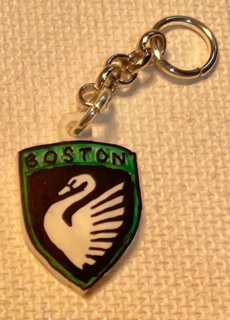 May include: A keychain with a shield-shaped emblem displaying the word "BOSTON" in green. The emblem features a black background and a white swan design. The keychain has a silver-coloured chain and ring.