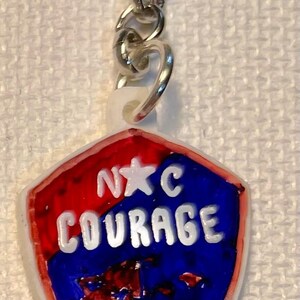 May include: A keychain featuring a shield-shaped pendant. The pendant is red and blue with the words "N☆C COURAGE" in white. A silver chain and ring are attached.