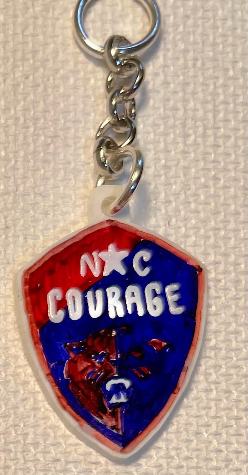 May include: A keychain featuring a shield-shaped pendant. The pendant is red and blue with the words "N☆C COURAGE" in white. A silver chain and ring are attached.