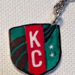 May include: Keychain with a shield design. The shield has the letters "KC" in white on a red central panel, with green and black detailing. Two white stars are on the right side. A silver chain and ring are attached.