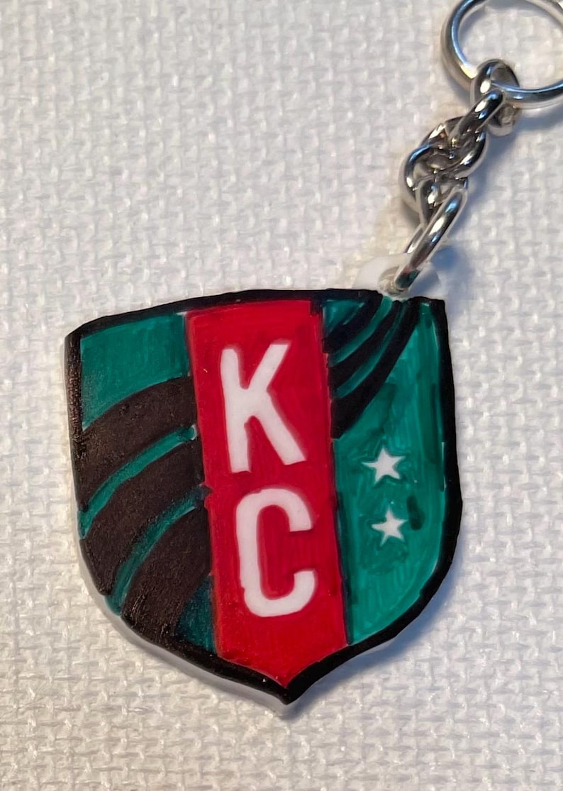 May include: Keychain with a shield design. The shield has the letters "KC" in white on a red central panel, with green and black detailing. Two white stars are on the right side. A silver chain and ring are attached.