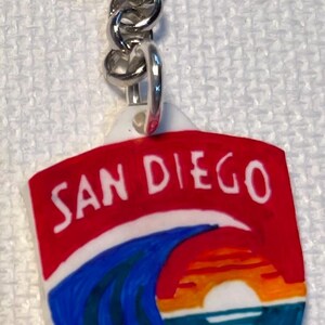 May include: A keychain with a shield-shaped pendant displaying the text "SAN DIEGO" in white on a red backdrop. Below the text is a depiction of a sunset over the ocean, with blue waves and an orange and yellow sky.