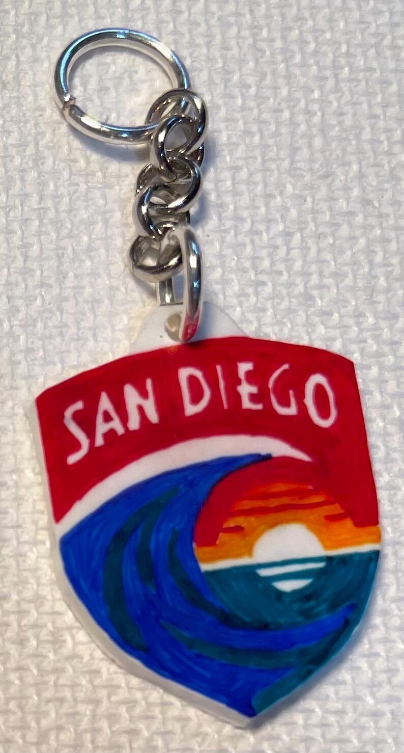 May include: A keychain with a shield-shaped pendant displaying the text "SAN DIEGO" in white on a red backdrop. Below the text is a depiction of a sunset over the ocean, with blue waves and an orange and yellow sky.