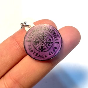 May include: A circular, purple Racing Louisville Football Club charm. The charm features black text and a fleur-de-lis design. It is attached to a silver-coloured chain.
