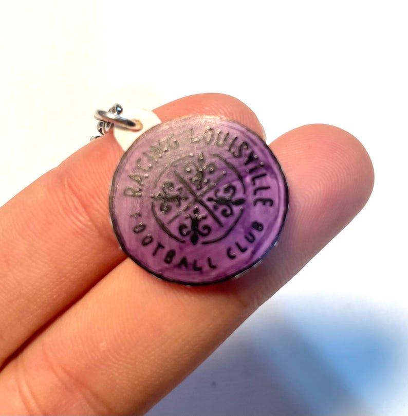 May include: A circular, purple Racing Louisville Football Club charm. The charm features black text and a fleur-de-lis design. It is attached to a silver-coloured chain.
