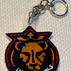 May include: A keychain with a silver ring and chain, featuring the words "UTAH ROYALS". The design includes a lion's head in orange and yellow, outlined in black and red. The keychain is a vibrant accessory.