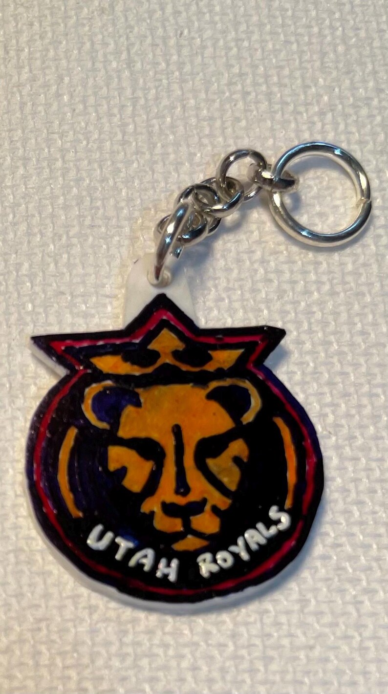 May include: A keychain with a silver ring and chain, featuring the words "UTAH ROYALS". The design includes a lion's head in orange and yellow, outlined in black and red. The keychain is a vibrant accessory.