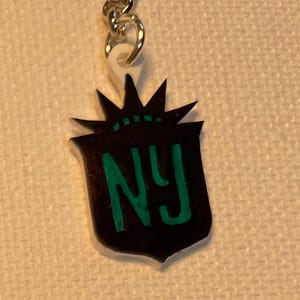 May include: A black and green NY keyring. The keyring features a shield-shaped emblem with the letters "NY" in green. The emblem is topped with a stylized crown design. The keyring is attached to a silver chain.