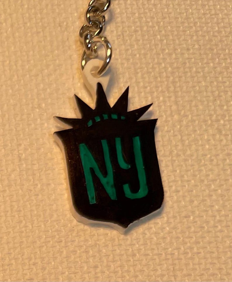 May include: A black and green NY keyring. The keyring features a shield-shaped emblem with the letters "NY" in green. The emblem is topped with a stylized crown design. The keyring is attached to a silver chain.