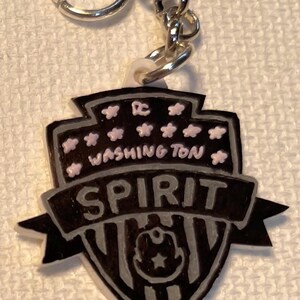 May include: A black and grey keychain featuring the words "WASHINGTON SPIRIT" and a team logo. The keychain has a silver chain and ring. The design includes pink stars and the letters "DC".