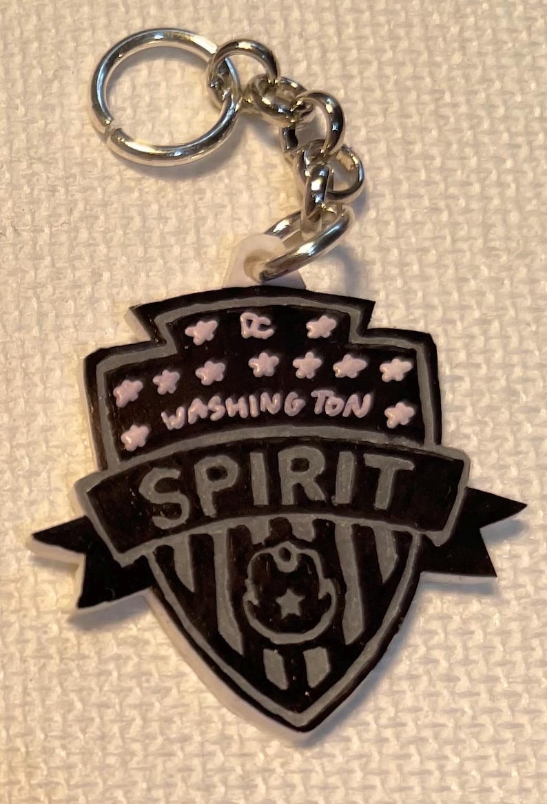 May include: A black and grey keychain featuring the words "WASHINGTON SPIRIT" and a team logo. The keychain has a silver chain and ring. The design includes pink stars and the letters "DC".