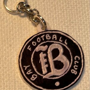 May include: A black and white keychain with the words "BAY FOOTBALL CLUB" around the edge. The centre features the letters "FB" in a stylized font. The keychain is attached to a silver metal ring.