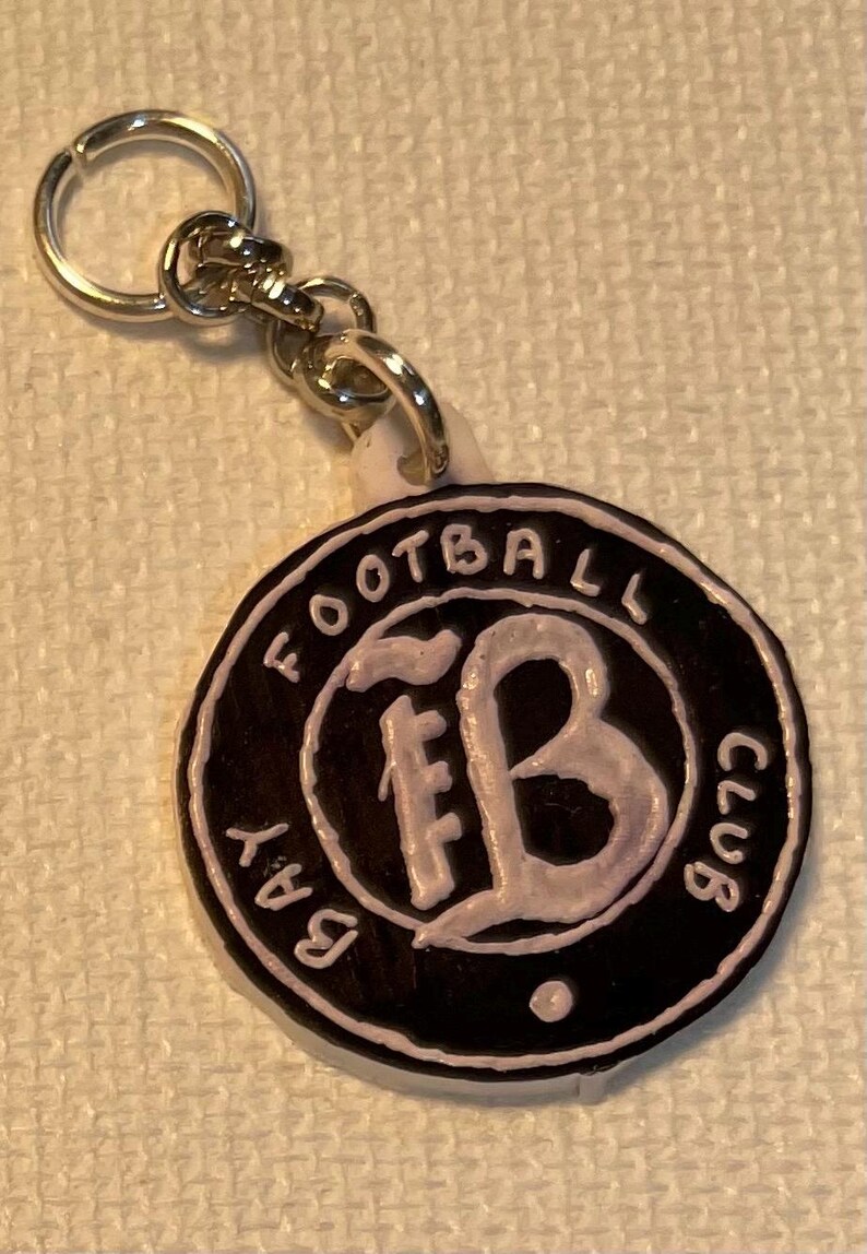 May include: A black and white keychain with the words "BAY FOOTBALL CLUB" around the edge. The centre features the letters "FB" in a stylized font. The keychain is attached to a silver metal ring.