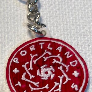 May include: A red and white Portland Thorns keyring. The round charm features the team name in white lettering around the edge, with a white design in the centre. The keyring has a silver chain and ring.