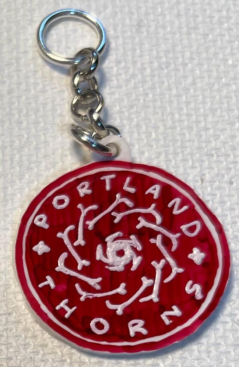 May include: A red and white Portland Thorns keyring. The round charm features the team name in white lettering around the edge, with a white design in the centre. The keyring has a silver chain and ring.